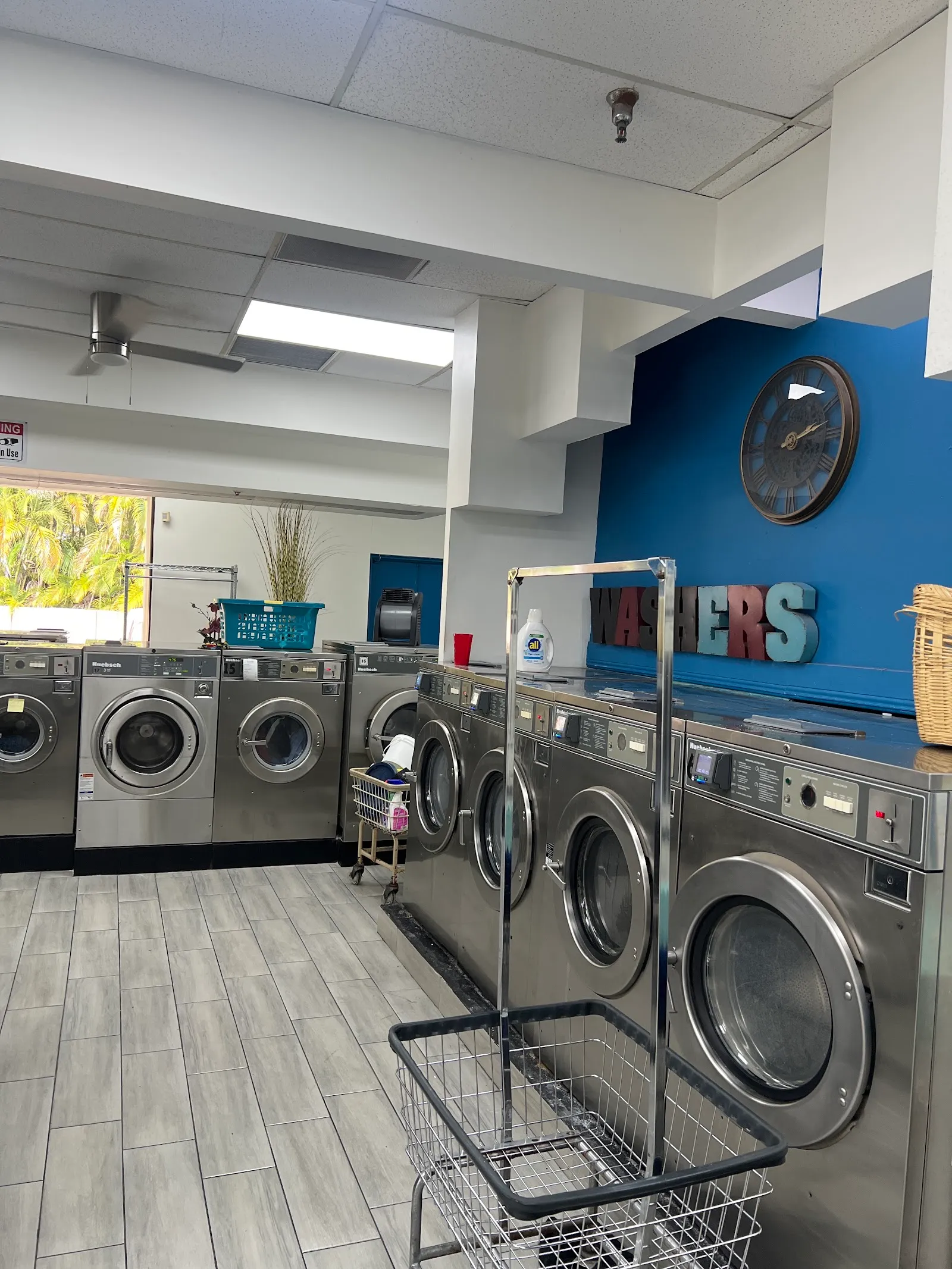 Interior view of the laundromat showing a row of stainless-steel front-load washing machines beneath a blue accent wall with a 'Washers' sign and wall clock.