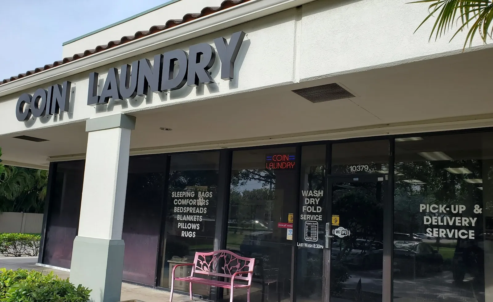 The Sunshine Kleen Coin Laundry storefront, with large 'Coin Laundry' signage, a pink bench out front, and window signs for wash-dry-fold and pickup & delivery service.